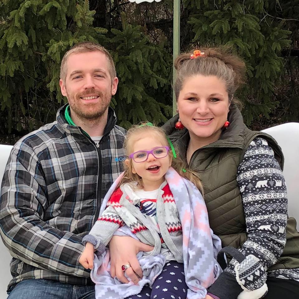 Dad, Mom, and young girl with purple glasses pose for a family photo outdoors during the Christmas season. 