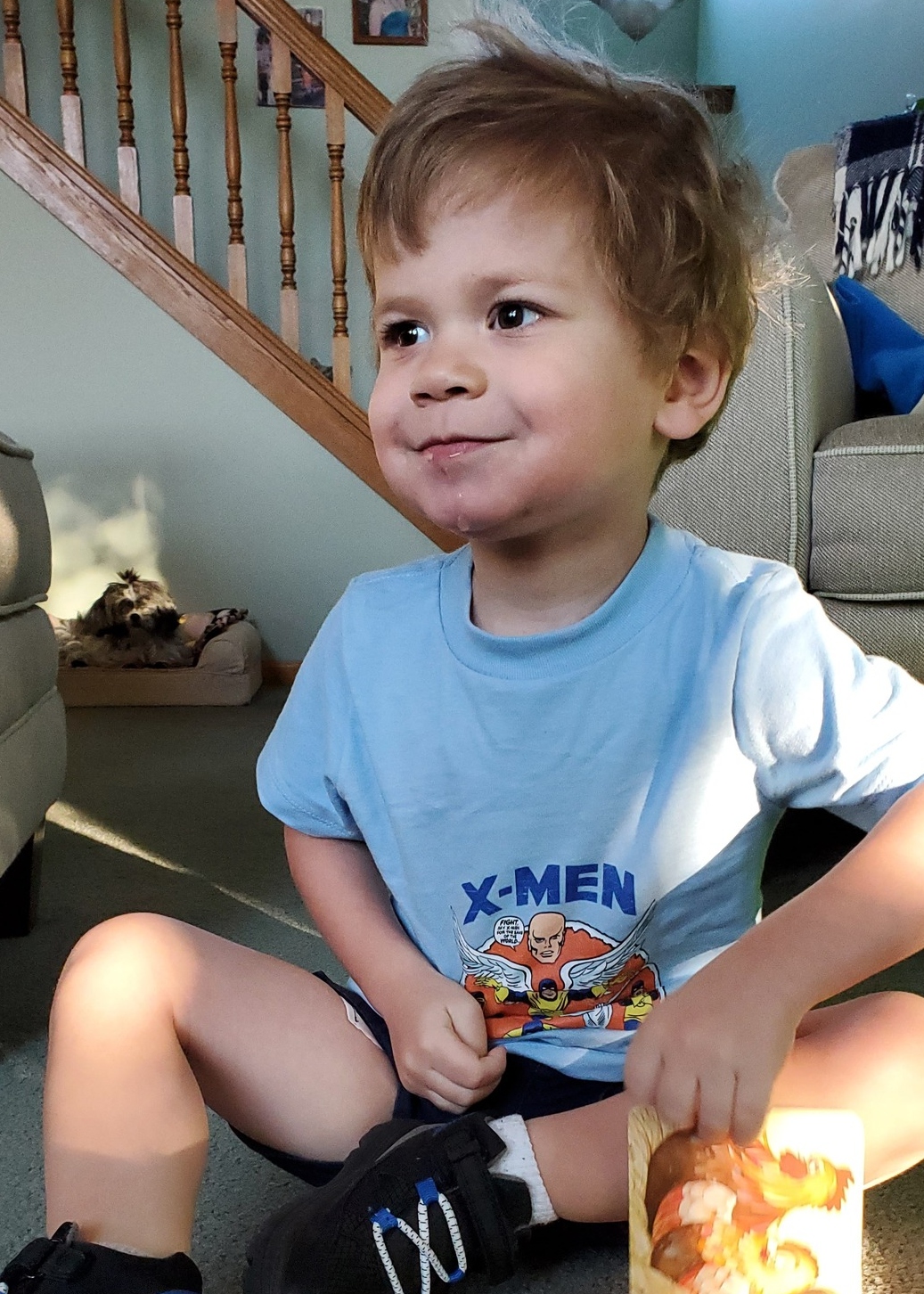 Little boy with blonde hair is sitting on the floor with a gentle smile wearing a blue X-Men t-shirt.