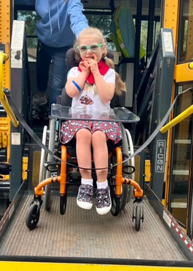 Little girl with green glasses on her way to school. Little girl is in a wheel chair and is getting lifted onto the bus with the wheel chair lift.