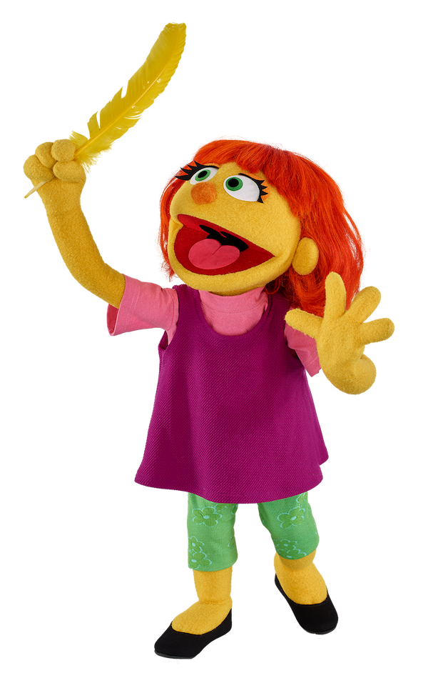 Julia, a puppet from Sesame Street, has yellow skin, red hair, and green eyes. She is wearing a purple dress with green leggings. She is holding a yellow feather with a big smile on her face.