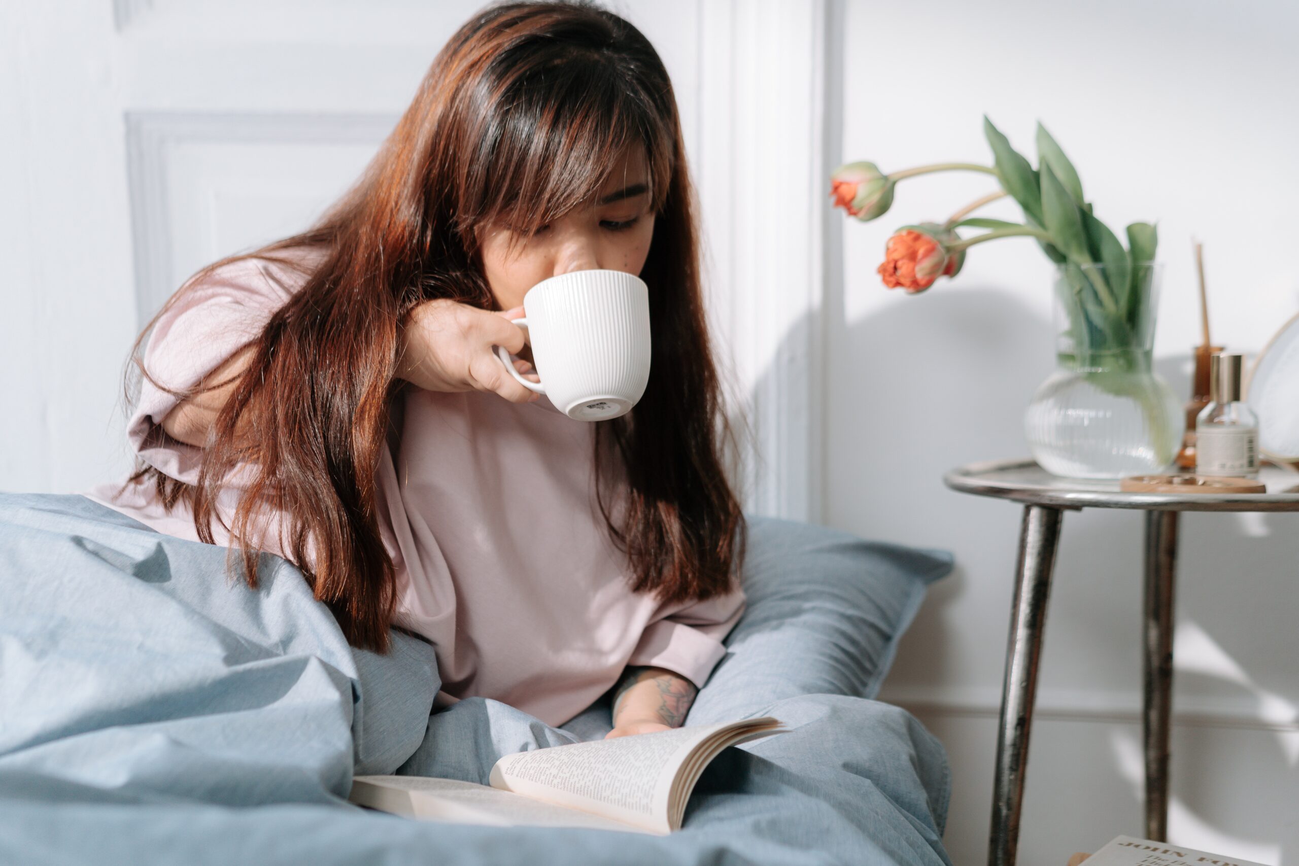 A woman sits in bed drinking out of a white mug while reading a book.